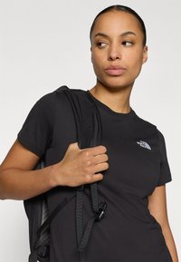 Μαύρο βαμβακερό μπλουζάκι με κοντά μανίκια, που φέρει λευκό λογότυπο στο αριστερό στήθος, φορεμένο με μαύρο λουρί σακιδίου που φαίνεται στον ώμο.