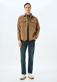 Vaaleanruskea villasekoitekangastakki kahdella etupussilla, kerroksittain ruutupaidan päällä, yhdistettynä farkkuihin ja valkoisiin lenkkareihin.