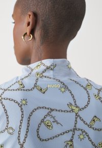 Světle modrá košile se vzorem řetězce a motivů v zelené barvě, se obrázky bot a hvězd. Zlaté logo "HUGO" umístěné na zádech.