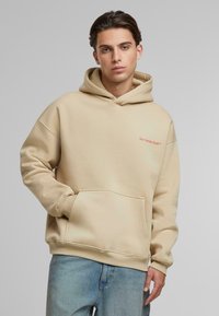 Beige fleece-hoodie med en frontlomme, ribbede manchetter og et logo i orange på venstre bryst. Bæres med lyseblå denimjeans.