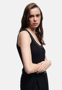Top sin mangas negro de canalé con escote redondeado y detalles de ribete plateado, combinado con pantalones negros de cintura alta y un cinturón negro.