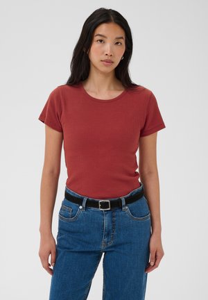 Joven mujer con cabello largo y negro, vestida con una camiseta de manga corta de color óxido y vaqueros azules con un cinturón negro, de pie frente a un fondo liso.