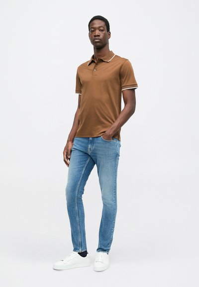 Camisa polo marrón con ribete blanco, combinada con jeans ajustados de color azul claro y zapatillas blancas; el modelo está de pie frente a un fondo neutro.