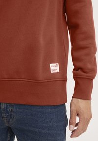 Roestkleurig sweatshirt met ribgebreide boorden en zoom, met een klein stoffen label aan de onderkant links. Draag het met donkerblauwe jeans.
