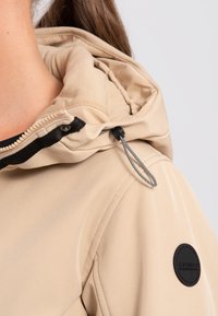 Beige hættejakke med tekstureret finish, der har en sort lynlås, justerbar snor og et rundt logopatch på ærmet.