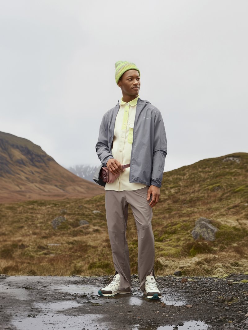 Mann in einer hellgrauen wasserdichten Jacke, gelbem Hemd, beigen Hosen und weißen Sneakers mit türkisfarbenen Akzenten, der im Freien steht.