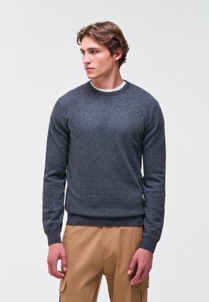 Jonge man met lichtbruin haar, die een grijze trui en beige cargobroek draagt, kijkt naar links tegen een effen lichte achtergrond.