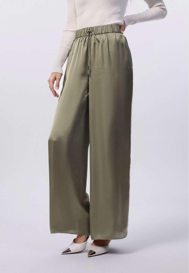 Pantalons larges en satin vert olive, avec une taille à cordon et des poches latérales. Associés à des talons blancs pointus.