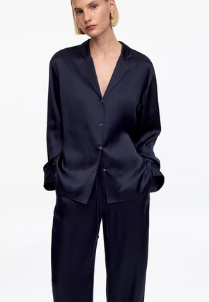 Žena s krátkymi blond vlasmi, ktorá má na sebe tmavo modrú saténovú košeľu na gombíky s dlhým rukávom a voľné nohavice v rovnakej farbe, ruky vreckách, neutrálne pozadie.