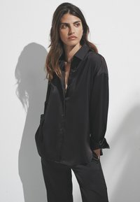 Kvinde med langt brunt hår iført løst sort satin skjorte og bukser, stående med hænderne i lommerne mod hvid baggrund.