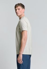 Lichtgrijze t-shirt met korte mouwen van zachte stof, met een ronde halslijn en een klein logo op de linkermouw. Draagt een navy broek.