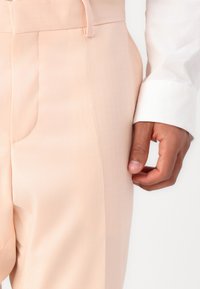 Lichtroze op maat gemaakte broeken met een gladde textuur, voorzien van een zijzak en een riemlus, gecombineerd met een witte shirtmanchet.