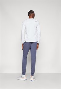 Weißes Langarmshirt mit Logo, graue Jogginghose und hellgraue Sportschuhe mit orangen Akzenten. Einfaches, lässiges Sportoutfit.