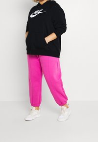 Person som bär en svart Nike-hoodie, ljusrosa sweatpants och vita sneakers, stående mot en enkel ljus bakgrund.