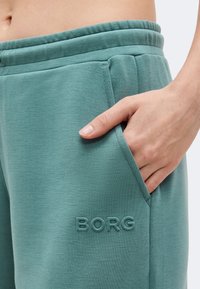 Κοντινή λήψη ενός χεριού που ακουμπάει στην πλαϊνή τσέπη ενός πετρόλ φόρμας με κεντημένο λογότυπο "BORG" κοντά στην τσέπη.