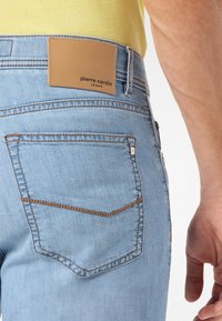 Ljust blå denimjeans bakficka med brun sömnad och en tan läderlapp märkt "pierre cardin JEANS," bärs med en gul skjorta.
