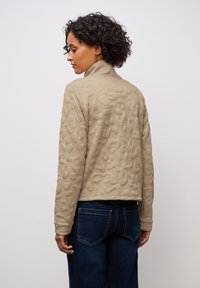 Beige vadderad jacka med upphöjt abstrakt mönster, hög krage och en kortad design, tillsammans med mörkblå vida jeans.