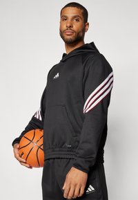 Sort træningshætte med hvide og røde striber på ærmerne, forlommerne og elastisk talje. Spiller holder en basketball.
