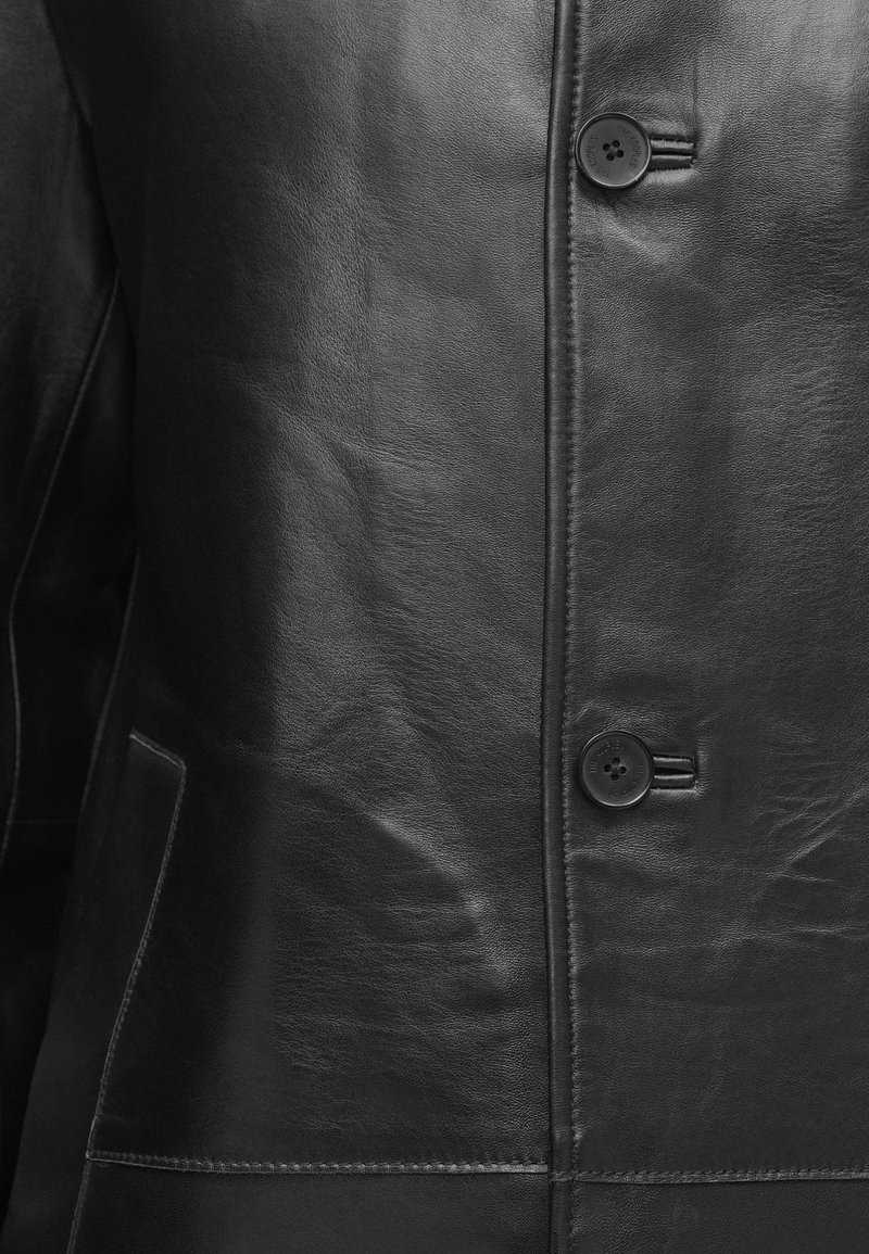 Gros plan sur une veste en cuir noir montrant une texture lisse, des détails de couture, deux boutons et une poche partielle sur le côté gauche.