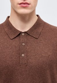 Νεαρός που φορεί καφέ πλεκτό πόλο μπλουζάκι με τρία κουμπιά και μικρό κεντημένο λογότυπο "GANT" στο στήθος.