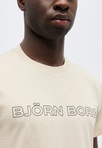 Κοντινό πλάνο ενός ατόμου που φοράει ανοιχτό μπεζ T-shirt με μαύρο περίγραμμα κειμένου «BJÖRN BORG» στο στήθος σε απλό φόντο.