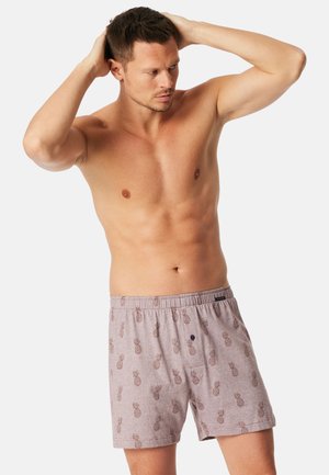Oberkörperfreier Mann mit kurzen Haaren, der hellgraue Boxershorts mit braunen Ananas-Mustern trägt, die Hände auf dem Kopf ruhend, schaut zur Seite.