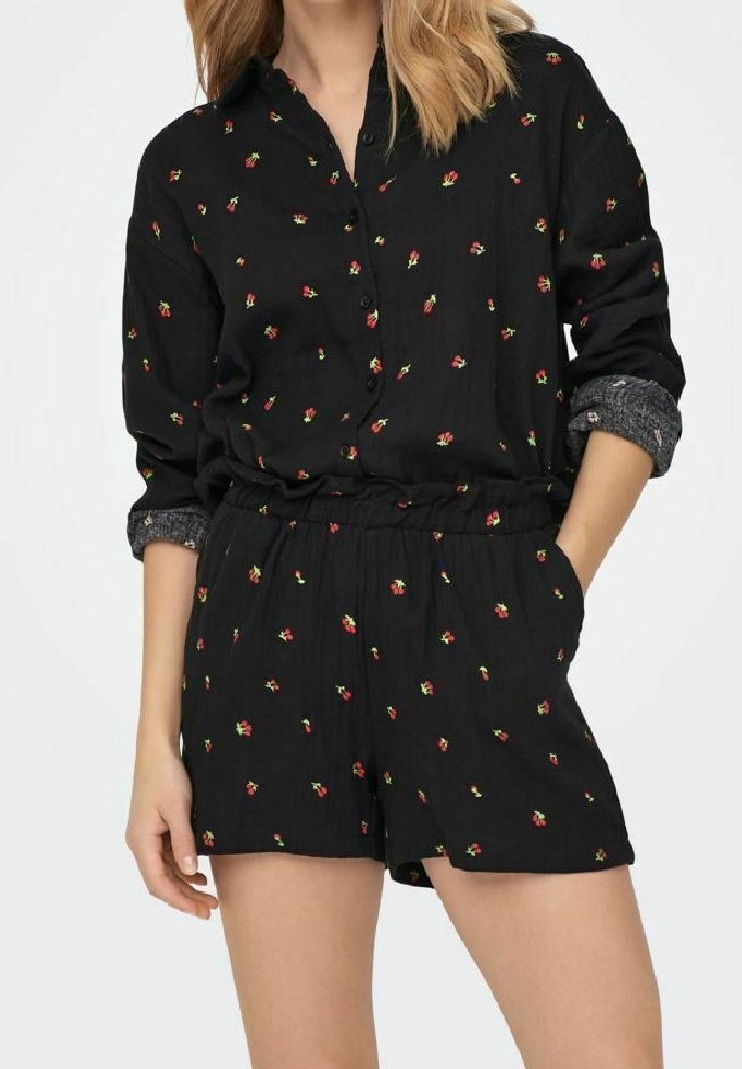 Femme portant une combinaison noire à manches longues boutonnée avec de petits motifs floraux rouges et jaunes, mains dans les poches, manches retroussées.