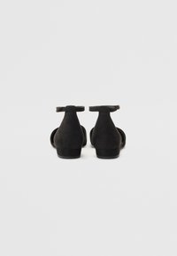 Zapatos de tacón de ante negro con correa en el tobillo y tacón bajo, diseño de espalda abierta y acentos de hardware mínimos. Textura suave con un acabado mate.