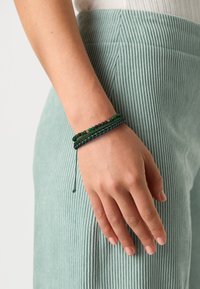 Zöld, fonott karkötő, különböző textúrájú gyöngyökkel, elegáns dizájnnal. Egy kézen viselve, texturált anyagon.