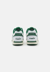 Valkoiset ja vihreät urheilukengät, joissa on teksturoitu kangas kantapää, rei'itetyt paneelit ja kumipohja, ja takana logo.