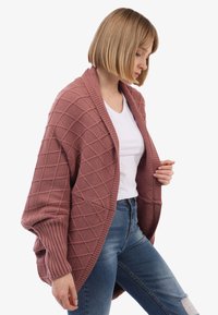 Frau mit kurzem blonden Haar, die eine strukturierte mauvefarbene Strickjacke über einem weißen T-Shirt und blauen, zerrissenen Jeans trägt und seitlich schaut.