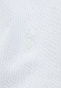 Hvid polo shirt stof med et struktureret mønster. Har et subtilt hvidt broderet hjortelogo på venstre side.