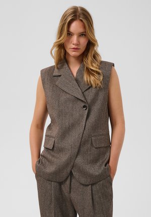 Femme blonde portant un blazer marron sans manches à chevrons et un pantalon assorti, debout les mains dans les poches devant un fond uni.