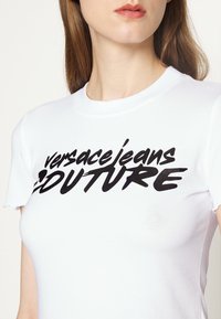 Vit t-shirt med kort ärm som har en framträdande svart logotyp med texten "Versace Jeans Couture" i ett fet, stiliserat typsnitt.