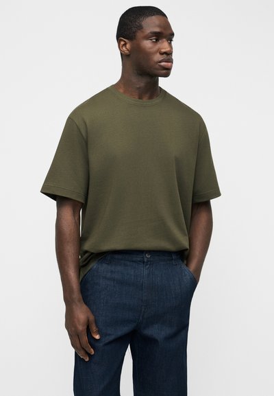 Άνδρας που φοράει ελαιζένιο πράσινο κοντομάνικο μπλουζάκι, κεντημένο μέσα σε σκουρομπλε τζιν, με τα χέρια στις τσέπες, κοιτάζει στο πλάι με φόντο λευκό.