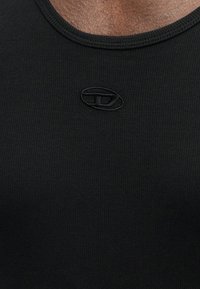 Primeiro plano de um tecido preto ribado com um pequeno logotipo oval costurado abaixo do decote em pele escura.