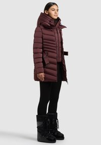 Bordo puhovka z visokim ovratnikom, vodoravnimi šivi in stranskimi zaponkami. Oblečena s črnimi legicami in usklajenimi snežnimi škornji.