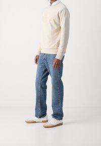 Ljusbeige sweatshirt med "BOSS"-logotyp, i kombination med blå jeans och vita sneakers med gummisula. Enkel rund halsringning och standard passform.