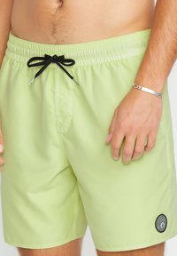 Pantaloni da bagno verde chiaro realizzati in tessuto morbido, con un laccio nero e una patch del logo circolare nella parte inferiore sinistra.