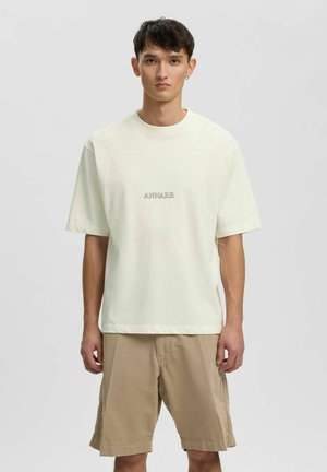 Ung mand iført en løs off-white T-shirt med "ANNARR" tekst og beige knælange shorts, stående mod en ensartet lys baggrund.