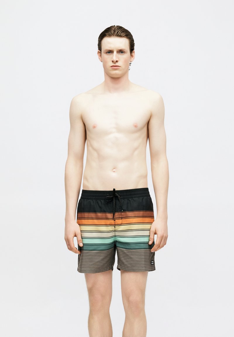 Νεαρός άνδρας με κοντά βρεγμένα μαλλιά, φορώντας πολύχρωμο ριγέ μαγιό, στέκεται μπροστά σε απλό λευκό φόντο, με ουδέτερη έκφραση.