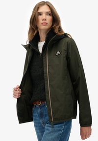 Ciemnozielona wodoodporna kurtka z zamkiem z przodu, regulowanym kapturem i haftowanym logo górskim. Noszona na czarnym swetrze w warkocze.