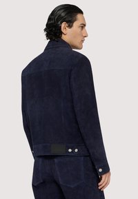 Námořnická semišová bunda se strukturovaným povrchem, s kovovými prvky v barvě stříbra, knoflíkovými detaily a zadním nášivkem. Jednoduchý design límce.