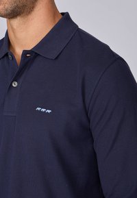 Polo shirt navy en tissu texturé avec col et deux boutons, arborant un petit motif brodé léger bleu de trois animaux.