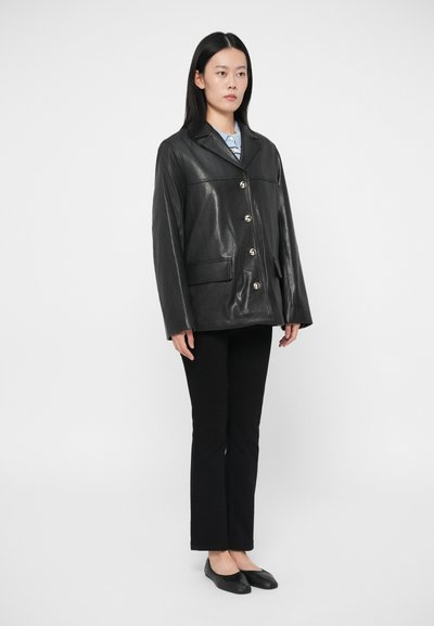 Γυναίκα που στέκεται φορώντας ένα μαύρο δερμάτινο σακάκι με κουμπιά, μαύρο παντελόνι, μαύρες μπαλαρίνες και ανοιχτό γαλάζιο πουκάμισο, μπροστά σε λευκό φόντο.