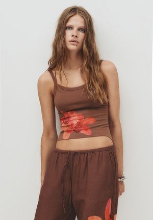 Jonge vrouw met lang golvend haar, gekleed in een bruine tanktop en een broek met grote rode bloemenprints, staand voor een effen witte achtergrond.