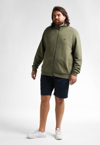 Chaqueta de cremallera verde oliva con patrón texturizado, cuello alto y logo. Combinada con pantalones cortos navy y zapatillas blancas.
