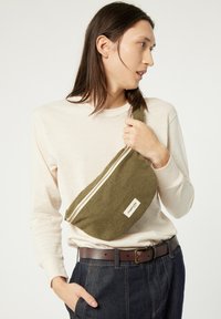 Pochette banane en tissu vert avec une fermeture zippée et une étiquette. Portée en bandoulière avec une chemise beige clair à manches longues et un pantalon en denim foncé.