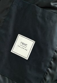 Tissu noir avec une étiquette carrée bordée de blanc, portant le texte « next » et « TAILORING ». Texture lisse, design minimaliste, sans motifs visibles.