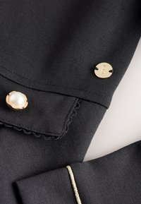 Tessuto nero con bordo smerlato, bottone in perla con rifinitura dorata, bottone rotondo dorato con la lettera "B" e sottile dettaglio di rifinitura dorata.